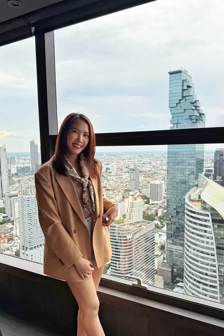 A woman in a camel blazer with the Bangkok skyline behind her