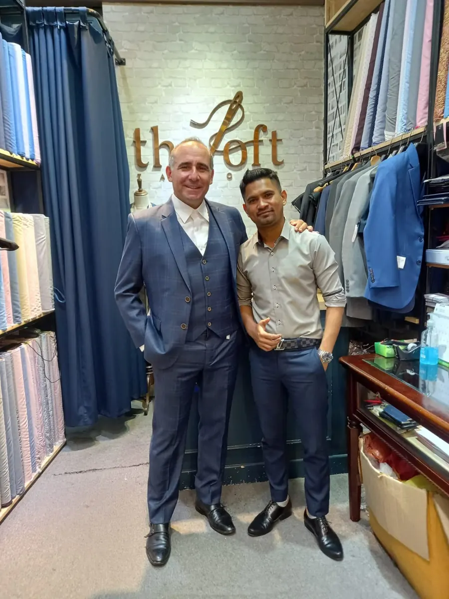 A customer in a navy three-piece suit with his tailor inside the shop